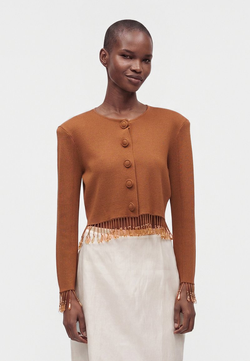 Γυναίκα που φοράει ένα σκουρόχρωμο cropped cardigan με κουμπιά, φουντάκια και λεπτομέρειες με χάντρες στις μανσέτες και το στρίφωμα, σε συνδυασμό με μια ανοιχτό μπεζ φούστα.
