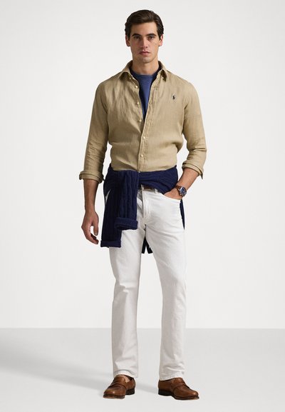 Beżowa lniana koszula, granatowy sweter związany w talii, niebieska koszulka, białe spodnie, brązowe eleganckie buty; postać stojąca na jasnym tle.