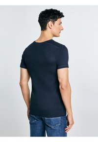 Hombre con una camisa ajustada de manga corta azul marino y jeans azules, de pie de perfil con la espalda parcialmente de cara a la cámara, sobre un fondo liso.