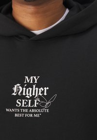Černá mikina s kapucí s výrazným bílým nápisem „MOJE Vyšší Já CHCE TO NEJLEPŠÍ PRO MĚ“ s dekorativní linií jako ozdobou.