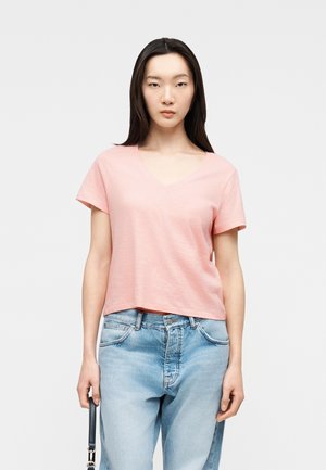 Jeune femme aux cheveux longs et raides, vêtue d'un t-shirt rose pâle à encolure en V et d'un jean bleu clair, tenant un sac à bandoulière noir sur un fond uni.