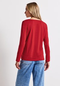 Långärmad topp med rundad halsringning i enfärgad röd tyg. Slät textur, figurnära design. Matchad med löst sittande ljusblå denimjeans.