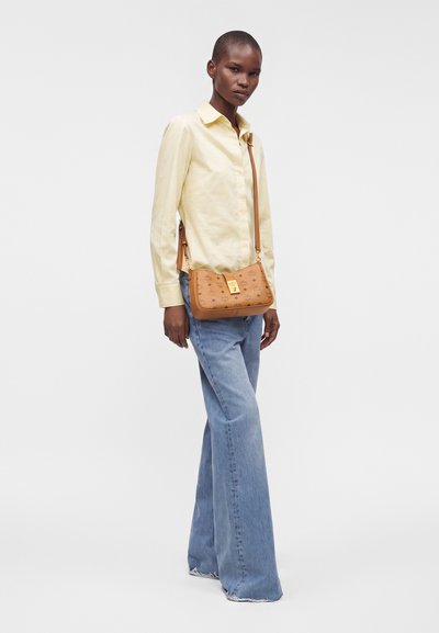 Femme portant une chemise à manches longues jaune clair, un jean bleu à jambes larges et un sac bandoulière beige avec une fermeture dorée.