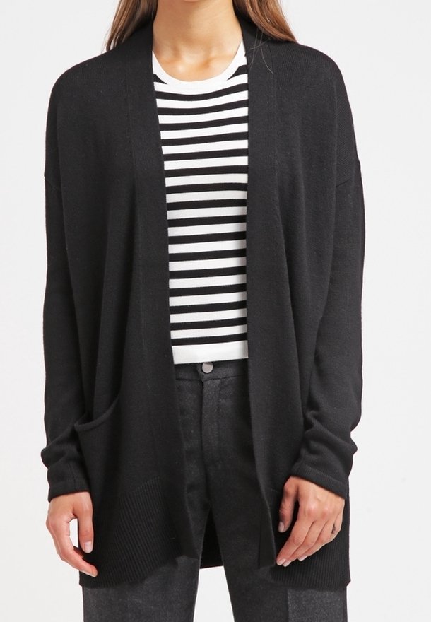 Femme portant un cardigan noir ouvert sur une chemise rayée noire et blanche et un pantalon gris foncé, debout, les mains détendues le long du corps.