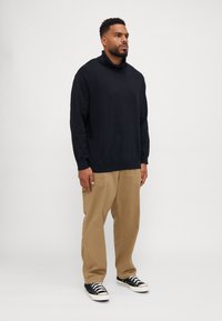 Schwarzer, langärmeliger Rollkragenpullover kombiniert mit beigen, lässig geschnittenen Hosen und schwarzen Sneakers. Einfaches Design mit umgeschlagenen Ärmeln und Saum.