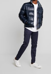 Doudoune marine à finition brillante, fermeture éclair, et poches latérales, portée au-dessus d'un t-shirt blanc et assortie de pantalons marins et de baskets blanches.