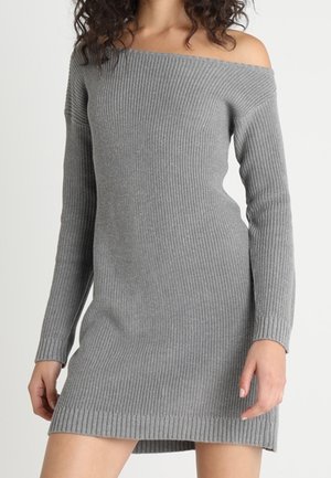 Vrouw draagt een grijze, geribbelde, off-shoulder gebreide jurk met lange mouwen, staande tegen een effen lichte achtergrond.