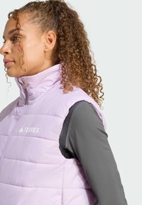 Hellviolette Steppweste mit hohem Kragen, mit Steppdesign und weißem Logo. Tragen darüber ein graues Langarmshirt, das einen lässigen Look zeigt.