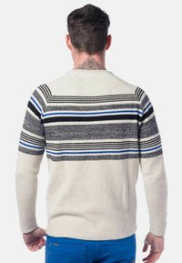 Homme portant un pull crème avec des rayures noires, grises et bleues sur les épaules et le haut du dos, associé à un pantalon bleu, debout dos à la caméra.