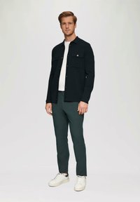 Donker button-up jack met twee borstzakken, gedragen over een wit t-shirt, gecombineerd met groene broek en witte sneakers. Glad materiaal.
