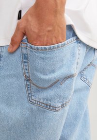 Džíny z lehce vypraného denimu s oranžovým prošíváním, s detailem zadní kapsy a rukou spočívající uvnitř kapsy.