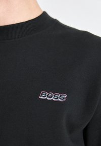 Felpa nera in tessuto morbido, con collo a costine e piccolo logo "BOSS" ricamato in rosa e bianco sul petto.