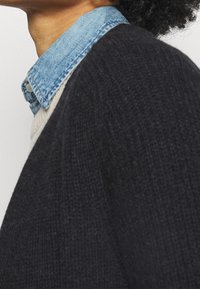 Tmavě modrý vlněný svetr s texturovaným pletením, přelitou přes světle modrou džínovou košili, která je viditelná na límci.