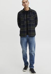 Flanellen overhemd in donker groen en marineblauw ruitpatroon, voorzien van twee borstzakken, gecombineerd met blauwe jeans en zwarte sneakers. Soepele, casual pasvorm.
