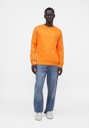 Mand iført en lys orange langærmet sweatshirt, lyseblå vidde jeans og hvide sneakers, stående mod en ensfarvet hvid baggrund.