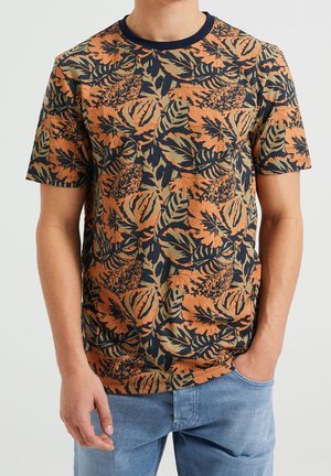 Mężczyzna ubrany w krótki rękaw, t-shirt z okrągłym dekoltem, ozdobiony pomarańczowym, czarnym i oliwkowozielonym wzorem tropikalnych liści, oraz jasnoniebieskie dżinsy.