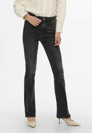 Kvinde iført sorte high-waist slim-fit jeans, cremefarvet blondebluse, beige spidse hæle og et gyldent armbånd på venstre håndled.