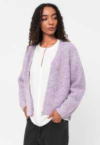 Lavendelfarbene Strickjacke mit strukturiertem Muster, offener Frontgestaltung und gerippten Bündchen. Getragen über einem weißen, knöpfbaren Top und kombiniert mit dunklen Hosen.
