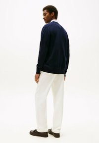 Dunkelblaues Strickpullover mit gerippten Bündchen und Saum, kombiniert mit weißen Hosen und braunen Loafers. Modell zeigt den Rücken und hebt das Design hervor.