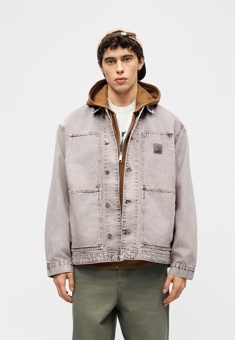 Jeune homme portant une casquette de baseball beige à l'envers, un sweat à capuche marron à fermeture éclair sous une veste en jean gris délavé avec de grandes poches, et un pantalon vert olive.