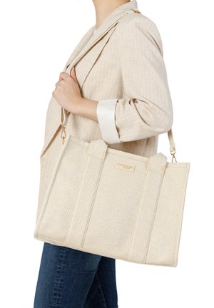 Frau in beigem Blazer und Jeans trägt eine große strukturierte cremefarbene Tragetasche mit Schulterriemen und aufgenähtem Logo.