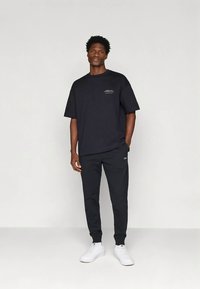 Muškarac stoji naslonjen na jednostavnu pozadinu, odjeven u crnu oversized majicu, crne trenirke i bijele tenisice, s jednom rukom u džepu.