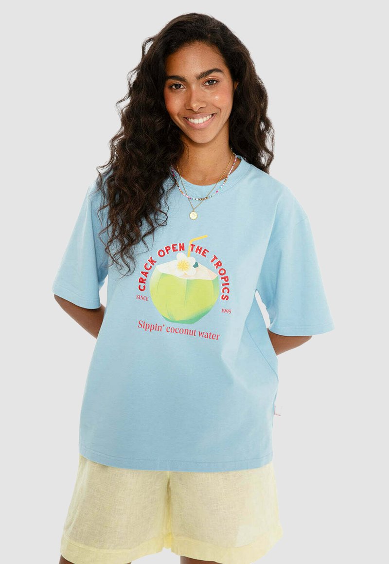 Světle modré tričko s krátkým rukávem má grafiku kokosu se brčkem a květinou, text "Otevři tropy" v červené barvě.