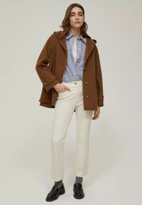 Cappotto di lana marrone con bottoni frontali, camicia bianca a righe blu, pantaloni bianchi a vita alta, mocassini neri e calze grigie.