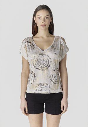 Kvinde iført en hvid og guldmønstret cold-shoulder top med sorte shorts, stående mod en ensartet lys baggrund.