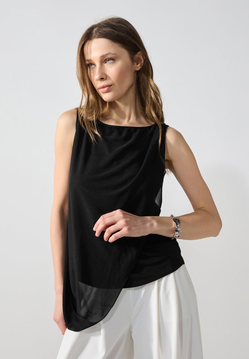 Black sleeveless top with draped front, smooth texture, and asymmetrical hem. Worn with white trousers and a silver bracelet.