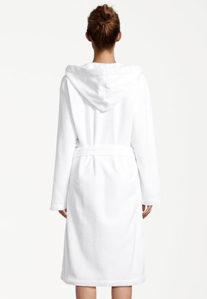 Frau mit zusammengebundenem Haar, die einen weißen Kapuzenbademantel trägt, der an der Taille gebunden ist, und mit dem Rücken zur Kamera vor einem schlichten Hintergrund steht.