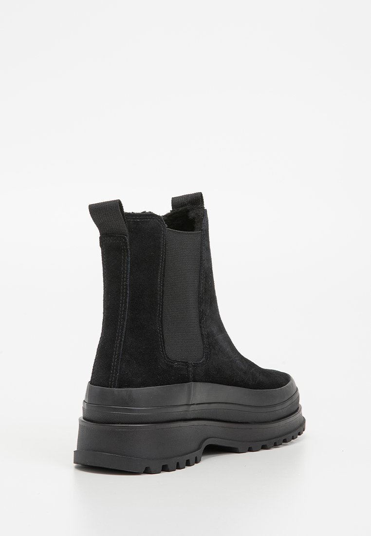 Botines de ante negro con suela de goma negra gruesa, paneles laterales elásticos y lengüetas en la parte superior para facilitar su uso. Se aprecian detalles de superficie texturizada.