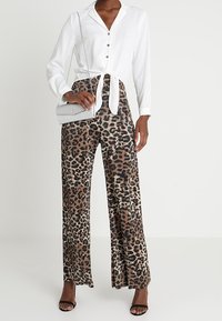 Chemise blanche à manches longues avec taille nouée, associée à un pantalon large imprimé léopard. Accessoirisée d'un sac bandoulière argenté et de talons noirs.