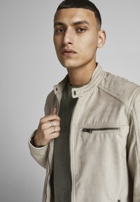 Bež suđena jakna s visokim ovratnikom, zatvorenim džepom i kviltiranim detaljima na ramenima, nosi se preko tamnozelene majice. Ruka modela se odmara blizu jakne.