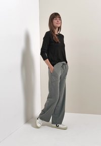 Femme aux longs cheveux bruns portant un haut noir à manches longues, un pantalon gris à jambes larges avec cordon et des baskets blanches, debout contre un mur uni.