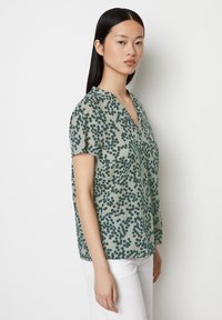 Kurzarmbluse mit V-Ausschnitt aus hellgrünem Stoff mit dunkelgrünem Punktmuster. Lockerer Schnitt und weiche Textur; glatt und ohne Verzierungen.