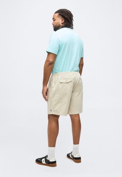 Άνδρας με κοτσίδες dreadlocks που φοράει ανοιχτό μπλε μπλουζάκι, μπεζ σορτς με πίσω τσέπες, άσπρες κάλτσες και ασπρόμαυρα αθλητικά παπούτσια, γυρισμένος προς τα πίσω.