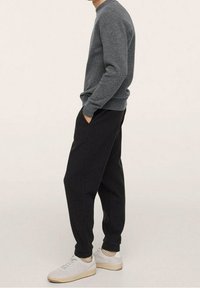 Grauer Strickpullover, lockere schwarze tapered Hose und weiße Sneakers mit beigen Sohlen. Seitenansicht vor einem neutralen Hintergrund.