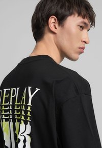 T-shirt in cotone nero con una grafica "REPLAY" in multicolore posizionata verticalmente sul retro. Collo a giro standard e maniche corte.