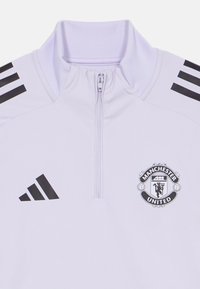 Bijela majica s patentnim zatvaračem, crnim Adidas prugama i logotipom Manchester Uniteda. Ima visoki ovratnik, glatku teksturu i moderan kroj.