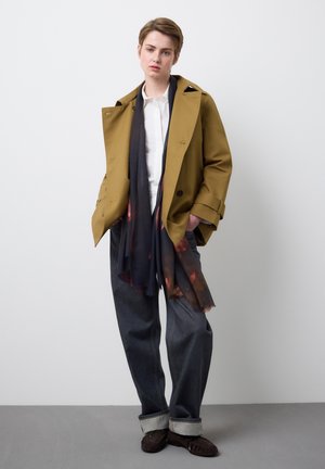 Giovane con cappotto color senape sopra camicia bianca, sciarpa floreale scura, jeans larghi con risvolti e mocassini marroni, in piedi contro una parete liscia.