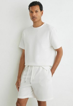 Man staat tegen een witte muur, draagt een volledig wit gestructureerd shirt met korte mouwen en bijpassende short met trekkoord.