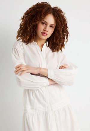 Junge Frau mit lockigem Haar in einem weißen Langarmkleid, die mit verschränkten Armen steht und selbstbewusst in die Kamera blickt.