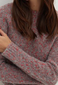 Close-up of a person wearing a gray fuzzy sweater with red speckles, arms crossed, showing long brown hair and partial face.