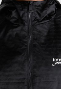 Svart jacka med en blank yta, med en framsida som har dragkedja, huva och ett upprepat logomönster i en subtil ton. Vitt varumärkeslogotyp på bröstet.