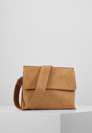 Tan corduroy crossbody tas met brede riem en klepsluiting, tentoongesteld op een witte sokkel tegen een effen grijze achtergrond.