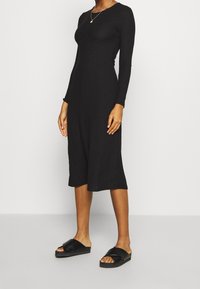 Svart ribbad midiklänning med långa ärmar och rund halsringning, tillsammans med svarta slidesandaler. Slät textur, figurnära siluett.