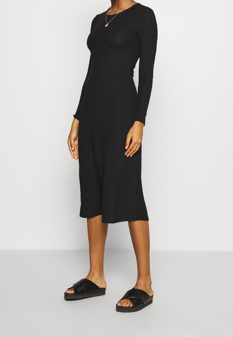 Svart ribbad midiklänning med långa ärmar och rund halsringning, tillsammans med svarta slidesandaler. Slät textur, figurnära siluett.