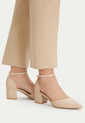 Mujer con zapatos de tacón ancho beige con correas en el tobillo, combinados con pantalones beige recortados, de pie sobre una superficie blanca.