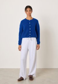 Jauna sieviete iekšpusē, tērpusies zilā kardiganā ar pogām, gaiši pelēnās platajiem bikšu kājslogiem un tumši brūnās šņorzābakās.